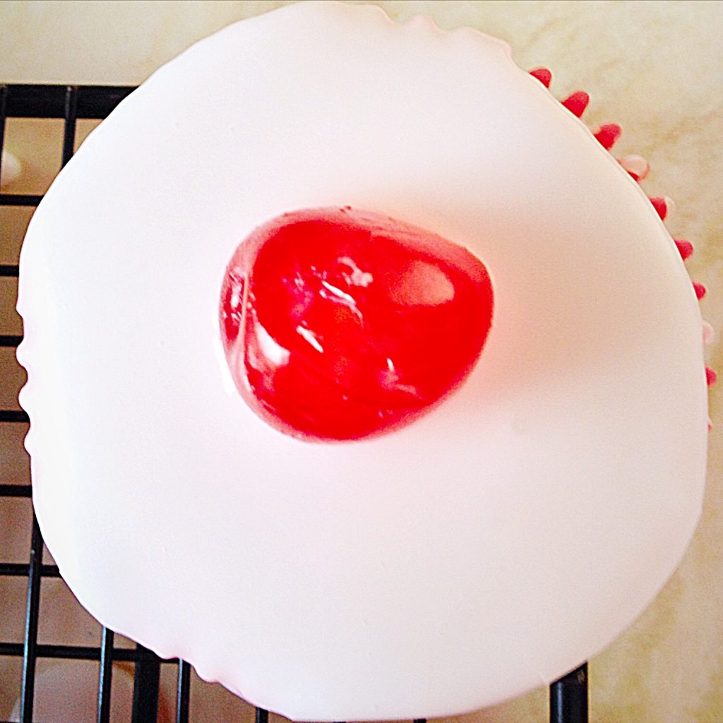 Bakewell Cupcakes topped with white icing and a red glacé cherry.