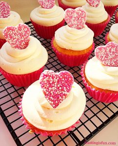Cupcakes in pink cupcake cases topped with piped white frosting and a single pink chocolate speckled heart.