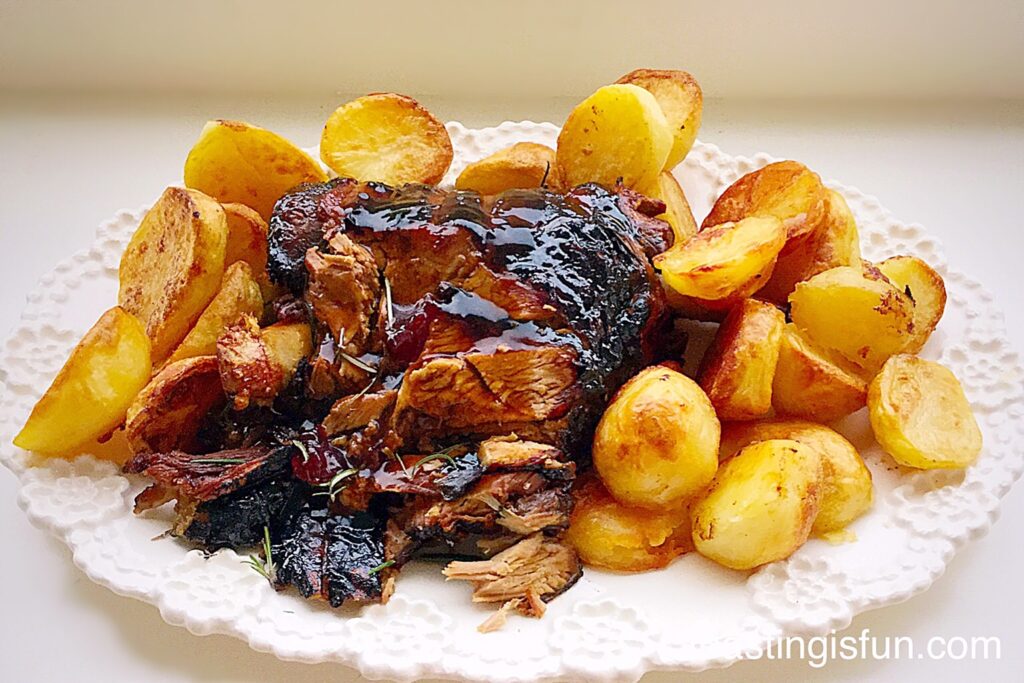 Roast leg of lamb sliced with a glaze poured over, surrounded by golden roast potatoes all on a white platter.
