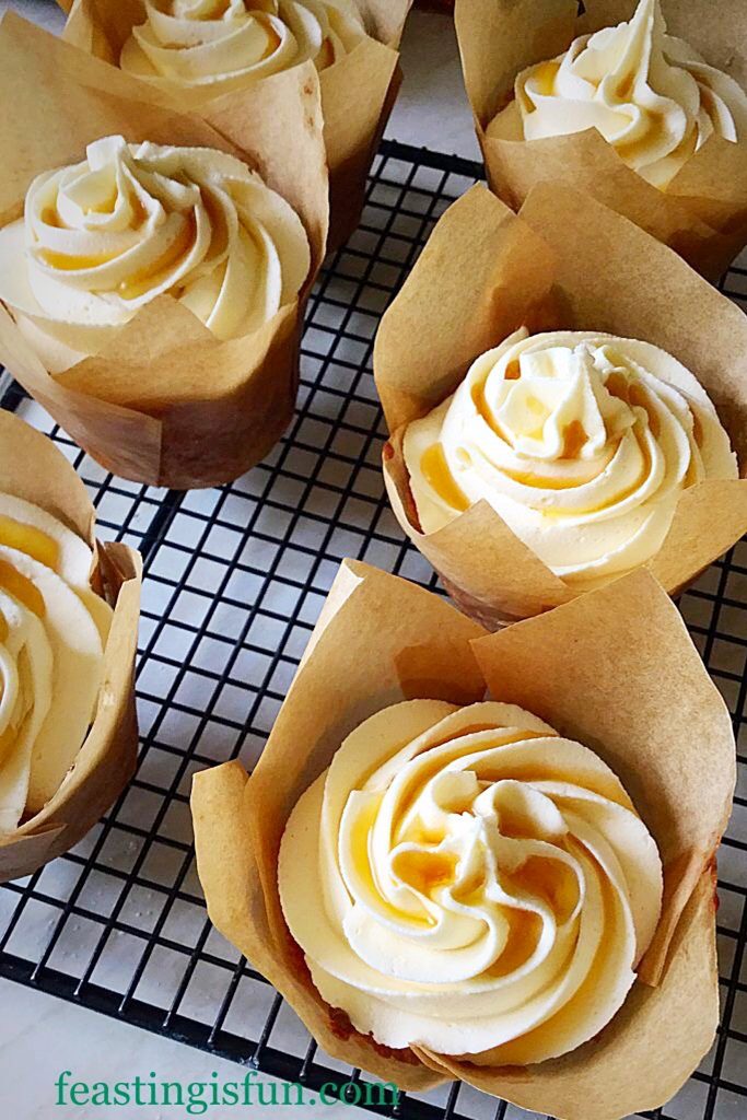 Freshly baked and frosted small cakes in caramel coloured tulip wrappers on a cooling rack.