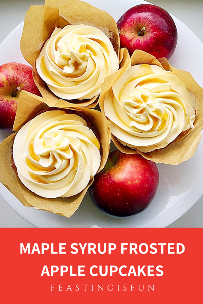Lightly spiced apple cupcakes in tulip wrappers, topped with creamy, piped frosting and a drizzle of maple syrup on a speckled white plate with apples.