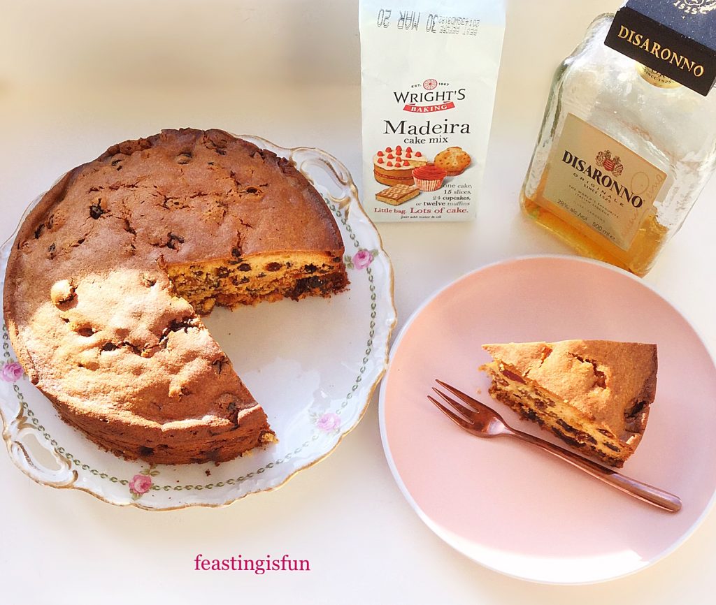 Cut amaretto light fruit cake on a cake plate with a slice of cake on a separate plate. A bottle of Disaronno liqueur and packet of cake mix.