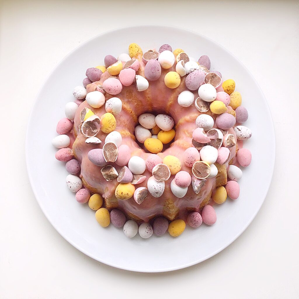 Looking down onto the top of a mini egg topped Bundt cake, surrounded by mini eggs on a white plate.