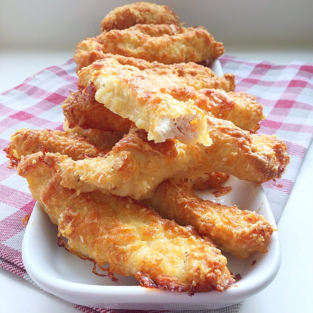 Golden and succulent chicken goujons stacked on a platter with a clear view of the inside of one strip.